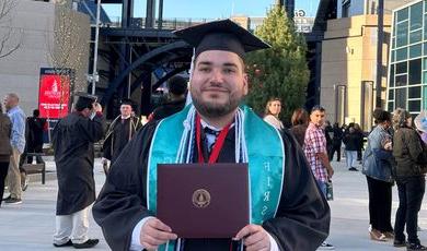 A man wearing graduation cap and gown holds a diploma in front of him 